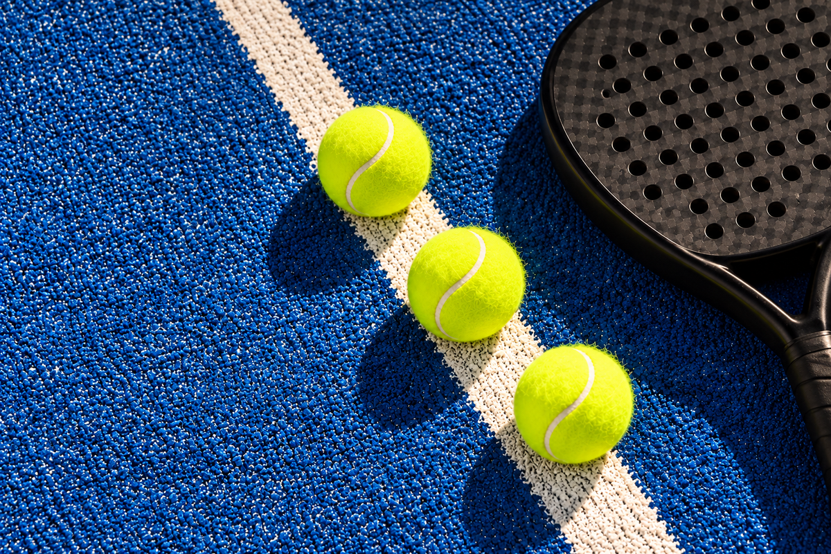 Three fresh yellow padel balls beside a racket on a blue court